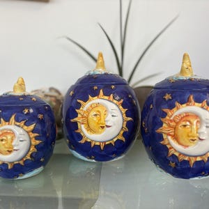 May include: Three spherical ceramic jars with lids, featuring a deep blue background decorated with gold stars. Each jar displays a raised, three-dimensional relief of a yellow sun and a white crescent moon face on the front surface.