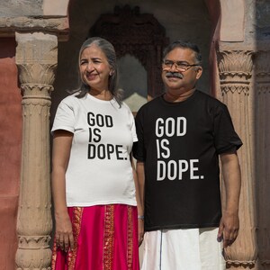 May include: A couple wearing matching white t-shirts with the text "God is dope." The woman is wearing a pink skirt and the man is wearing white pants. They are standing in front of a stone archway.