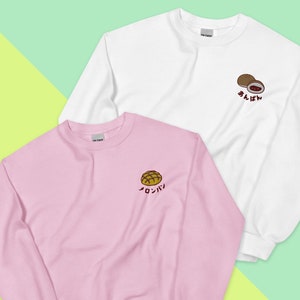 May include: Two sweatshirts with Japanese text and embroidered images of bread. The pink sweatshirt has a brown bread roll with the text "メロンパン" and the white sweatshirt has two brown bread rolls with the text "あんぱん".