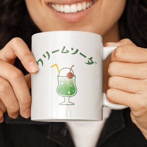 Puede incluir: Una taza de cerámica blanca con una ilustración verde y blanca de un flotador de soda cremosa japonesa. La taza tiene el texto japonés "クリームソーダ" impreso en ella.