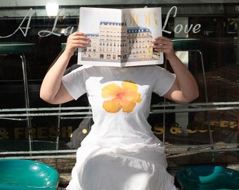 以下が含まれることがあります： 白いTシャツに黄色のハイビスカスの花柄がプリントされた白いスカートを履いた女性が窓の前に座っています。彼女は顔の前に雑誌を持っています。雑誌の表紙には「ADOR」と書かれています。