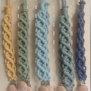 May include: Five macrame dummy clips in shades of yellow, green, light blue, sage green, and navy blue. Each clip has a wooden bead and a metal clip. The clips are displayed on a wooden pegboard.