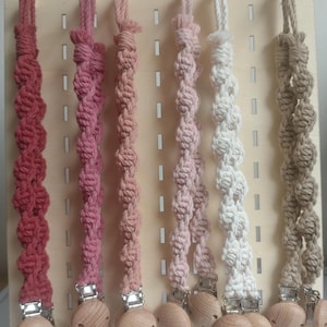 May include: A selection of macrame pacifier clips in shades of red, pink, and beige. Each clip has a wooden bead and a metal clasp. The clips are displayed on a wooden pegboard.