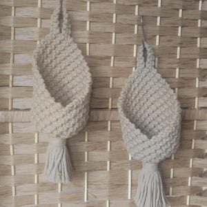 Peut inclure: Deux suspensions pour plantes en macramé beige, avec une texture tissée et des détails de pompons, suspendues sur un fond tissé. Ces supports décoratifs sont conçus pour suspendre des plantes.