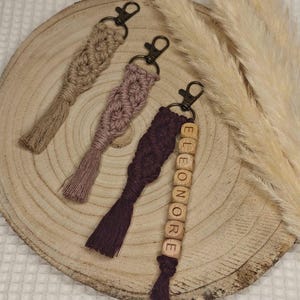 Puede incluir: Tres llaveros de macramé hechos a mano con borlas. Un llavero es beige, otro es rosa y el último es morado. El llavero morado tiene cuentas de madera con el nombre "Eleanor" escrito.
