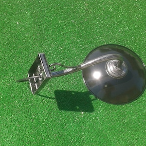 Op de afbeelding: Een zwarte metalen wandlamp met een gebogen arm en een ronde, zwarte kap. De lamp is gemonteerd op een groene ondergrond.