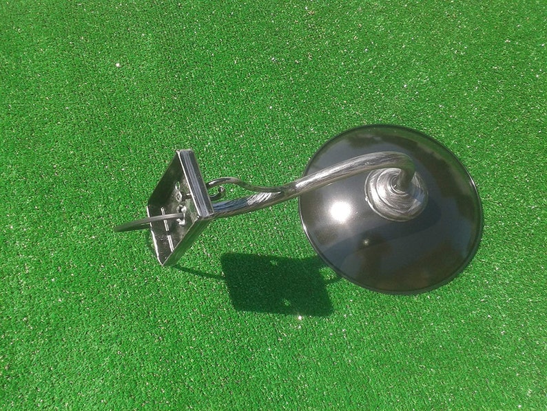 Op de afbeelding: Een zwarte metalen wandlamp met een gebogen arm en een ronde, zwarte kap. De lamp is gemonteerd op een groene ondergrond.