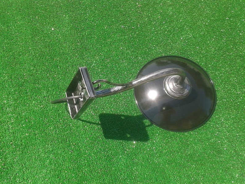 Op de afbeelding: Een zwarte metalen wandlamp met een gebogen arm en een ronde, zwarte kap. De lamp is gemonteerd op een groene ondergrond.