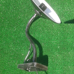 Op de afbeelding: Een zilveren metalen wandlamp met een gebogen arm en een ronde, platte kap. De lamp heeft een zwart elektriciteitskoord en een zwarte metalen basis.