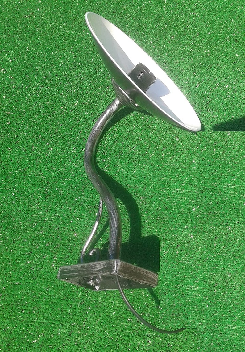 Op de afbeelding: Een zilveren metalen wandlamp met een gebogen arm en een ronde, platte kap. De lamp heeft een zwart elektriciteitskoord en een zwarte metalen basis.