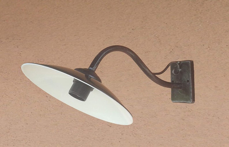 Op de afbeelding: Een witte metalen buitenwandlamp met een gebogen arm en een witte kap. De lamp is gemonteerd op een bruine muur.