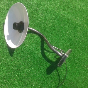 Op de afbeelding: Een witte metalen lamp met een gebogen arm en een witte kap. De lamp is aan de muur gemonteerd en heeft een zwart snoer.