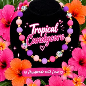 May include: A beaded necklace featuring round matte beads in shades of pink, purple, and white with floral patterns, interspersed with small metallic gold beads. The text "Tropical Candycore" and "Handmade with Love" is visible.