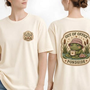 May include: A cream-colored t-shirt featuring a small graphic on the front chest with the text The Creature Club and a larger graphic on the back showing a frog wearing a floral bucket hat, holding a mug, with the text Out of Office Pondside.