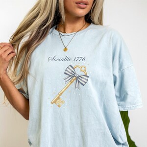 May include: Light blue t-shirt with the text "Socialite 1776" above a gold key design with a blue and white striped bow. The shirt has short sleeves and a relaxed fit.