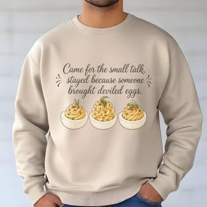 May include: A beige crewneck sweatshirt featuring the dark brown text "Came for the small talk, stayed because someone brought deviled eggs." Below the text are three illustrations of deviled eggs, each topped with different green herbs.