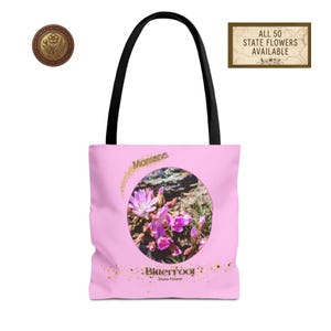 May include: Pink tote bag featuring a circular image of Bitterroot flowers, the Montana state flower. The bag has black handles and the text "Montana" and "Bitterroot State Flower" are printed on the front. A gold seal and a sign that says "All 50 State Flowers Available" are also visible.