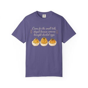 May include: A purple short-sleeve t-shirt featuring the text "Came for the small talk, stayed because someone brought deviled eggs." and a graphic illustration of three deviled eggs topped with green herbs arranged in a horizontal row.