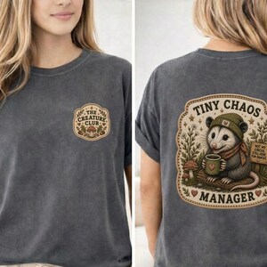 May include: A charcoal gray t-shirt featuring a small logo on the front chest that reads The Creature Club with a mushroom graphic. The back displays a large illustration of an opossum wearing a beanie and scarf, holding a mug, with the text Tiny Chaos Manager.
