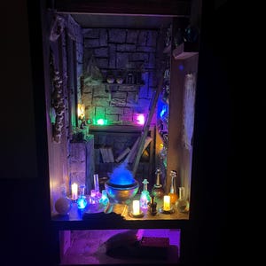 Könnte beinhalten: Miniatur-Diorama einer Hexenwerkstatt, beleuchtet mit farbigen LED-Lichtern. Die Szene umfasst einen Kessel mit blauem Rauch, Zaubertränke, Kerzen und eine Steinmauer. Das Gesamtthema ist Fantasy und Magie.