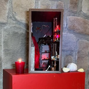 May include: A miniature diorama in a wooden frame, featuring a dark interior with a fireplace, a wand, and a red feather. A red candle burns on a red surface, with garlic cloves and a bulb nearby. The scene is set against a stone wall.