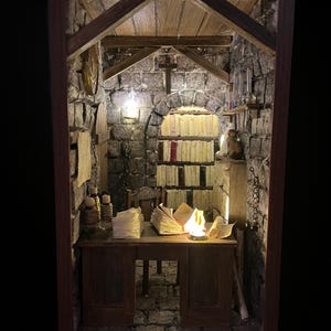 Könnte beinhalten: Miniatur-Diorama eines mittelalterlichen Arbeitszimmers mit Steinmauern, Holzbalken und einem Schreibtisch. Auf dem Schreibtisch liegen ein offenes Buch, eine brennende Kerze und Papierstapel. Bücherregale befinden sich im Hintergrund, beleuchtet von warmem Licht.