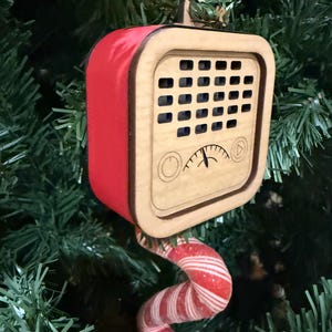 May include: A wooden Christmas tree ornament shaped like a vintage radio with a red side panel and a candy cane-patterned base. The front features a speaker grille, a circular dial with a needle, and two circular buttons with play and power icons.