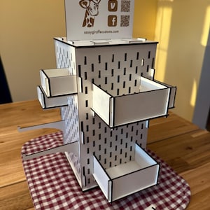 Peut inclure: Un présentoir blanc avec des bordures noires et plusieurs petits tiroirs. Le présentoir porte un logo de girafe et le texte "Sassy Giraffe Customs". Il est posé sur un tissu à carreaux rouges et blancs.