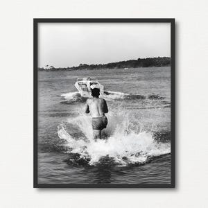 May include: A black and white photograph of a person waterskiing behind a boat on a lake. The person is wearing a bathing suit and is being pulled by a boat that is out of focus in the background.