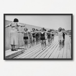 May include: A black and white photograph of a group of girls in swimsuits standing on a wooden pier. They are all wearing swim caps and are holding their arms out to the sides. A woman in a skirt is standing at the end of the pier, facing the girls.