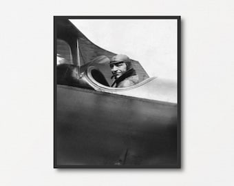 Peut inclure: Tirage encadré en noir et blanc d'un avion biplan d'époque et de son pilote. Le pilote, portant un casque et une veste en cuir, est assis dans le cockpit ouvert. L'aile de l'avion est visible, avec un ciel nuageux en arrière-plan.