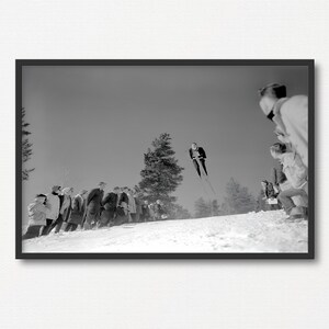 May include: A black and white photograph of a skier in mid-air, jumping off a ski jump. A crowd of people watches from below, standing on a snowy slope.
