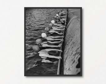 May include: Black and white photograph of a group of women in swimsuits and swim caps lined up in a row in a pool. The women are all facing the camera and have their arms outstretched. The water is rippling around them.