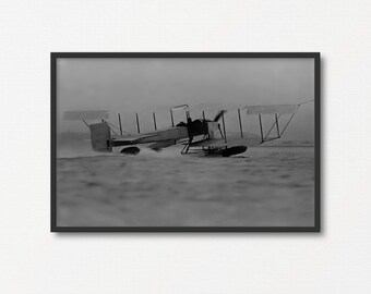Peut inclure: Une photographie en noir et blanc d'un hydravion vintage décollant de l'eau. L'avion est en plein vol, ses roues quittant à peine la surface de l'eau. L'avion est entouré d'une gerbe d'eau.