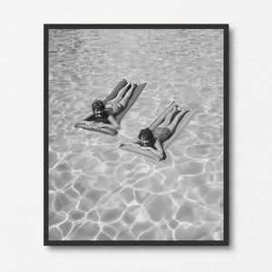 May include: Black and white framed print of two people relaxing on inflatable rafts in a pool. The water's surface reflects sunlight, creating a shimmering effect. The people are wearing swimsuits and enjoying the summer day.