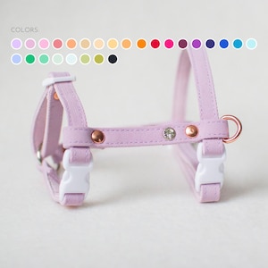 May include: A light purple pet harness featuring white plastic side-release buckles, copper-toned metal rivets, and a single clear rhinestone accent. The harness includes a copper-toned D-ring for leash attachment and a color chart labeled COLORS.