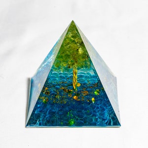 Peut inclure: Une pyramide en verre transparent avec un arbre en cristal vert à l'intérieur. La base de la pyramide est remplie de paillettes bleues et dorées.