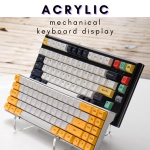 Könnte beinhalten: Zwei mechanische Tastaturen, die auf einem klaren Acryl-Ständer ausgestellt sind. Die obere Tastatur hat schwarze Tasten mit weißen Beschriftungen und gelben Akzenten. Die untere Tastatur hat weiße Tasten mit schwarzen Beschriftungen und gelben Akzenten. Die Tastaturen befinden sich auf einer Holzoberfläche.