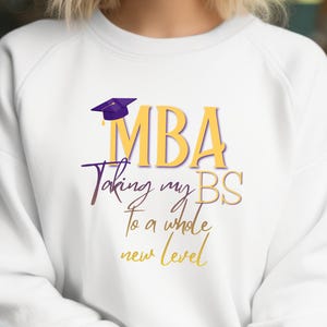 May include: White sweatshirt with the text "MBA Taking my BS to a whole new level" in gold and purple. The design includes a graduation cap. The sweatshirt is a casual style.