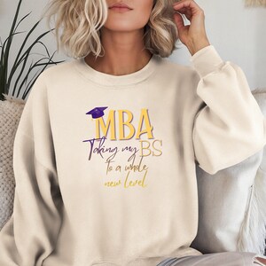 May include: Cream-colored sweatshirt with the text "MBA Taking my BS to a whole new level" in purple and gold lettering. A purple graduation cap graphic is above the word "MBA". The sweatshirt has long sleeves and a crew neck.