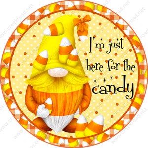 May include: A round, orange and white checkered background with a yellow gnome wearing a candy corn hat. The gnome is holding a candy corn and the text "I'm just here for the candy" is written in black.