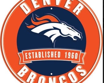 Puede incluir: Un letrero redondo de metal naranja y azul con las palabras "DENVER BRONCOS" en letras blancas. El letrero también presenta un logotipo de cabeza de caballo blanco con llamas naranjas y el texto "ESTABLISHED 1960".