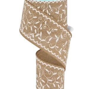 May include: A roll of decorative ribbon with a natural burlap texture. The ribbon features a white, scalloped edge and a repeating floral pattern. The spool has a teal and white label that reads "RG Ribbon".