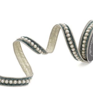 May include: A spool of dark teal velvet ribbon featuring a central row of small white faux pearls bordered by metallic silver trim. The ribbon is partially unrolled, displaying a textured light gold underside and a consistent pearl pattern.