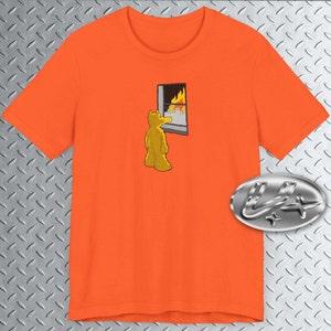 May include: Orange t-shirt with a cartoon illustration of a yellow bear looking at a window with flames inside. The bear is standing on a gray surface.