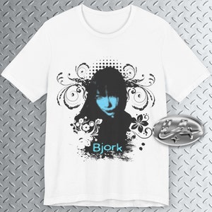 May include: White t-shirt with a black and white graphic design featuring a woman's face with blue eyes and a floral design. The text "Bjork" is printed in blue on the shirt.
