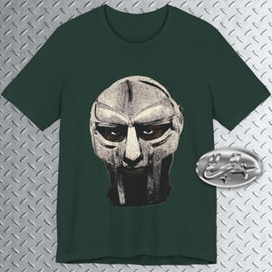 May include: Dark green t-shirt featuring a detailed illustration of a mask. The mask is primarily beige with black shading, creating a textured appearance. The shirt has a classic crew neck design and short sleeves. A silver-colored emblem is on the right side.