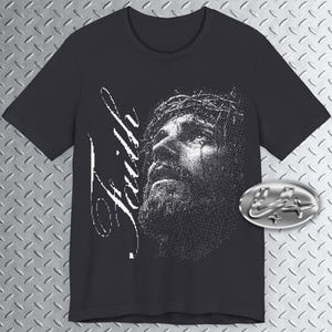 Puede incluir: Camiseta negra con una imagen en blanco y negro de un hombre con barba y corona de espinas. La palabra "Faith" está escrita en blanco a la izquierda. Un emblema ovalado plateado está en el lado derecho.