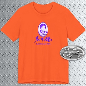 May include: Orange t-shirt with a purple image of a person's face in an oval frame. The text "Lao Gan Ma" is printed below the image in purple.
