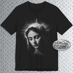 May include: Black t-shirt featuring a detailed, dot-style illustration of a woman with a halo. The image is in grayscale, with the woman's face and halo rendered in white against a black background. A silver-colored emblem is on the right side of the shirt.